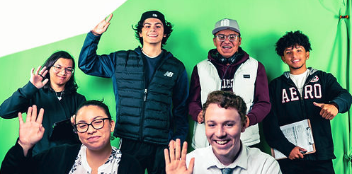 Group of students pose and wave in front of a green screen, with several standing and seated together in a studio-style setting.