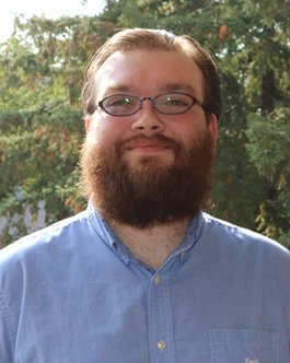 Professional headshot of a man with glasses and a full beard wearing a light blue button-down shirt outdoors.