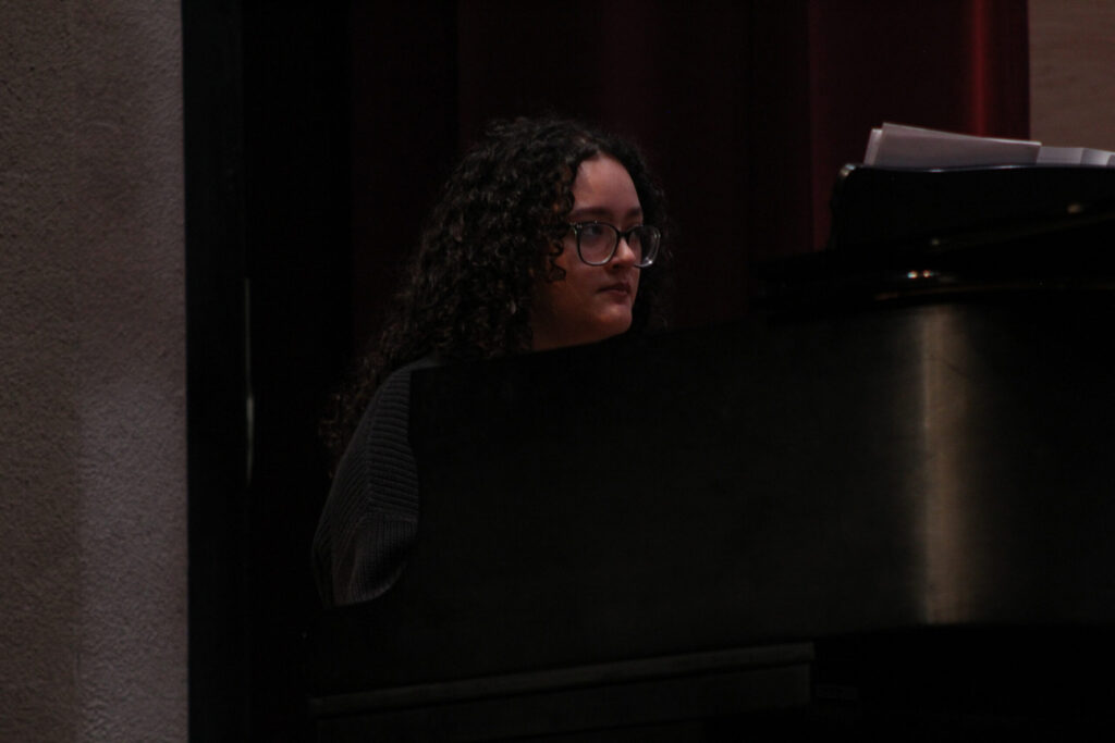 Woman with curly dark hair and glasses seated at a piano in a dimly lit performance space.